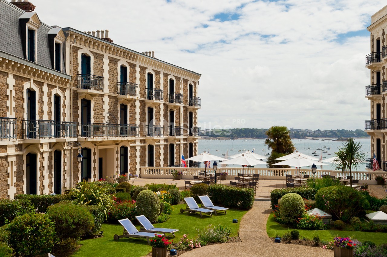 Cour intérieure d'un hôtel avec jardin aménagé, transats, parasols et vue sur la mer avec des voiliers au loin.
