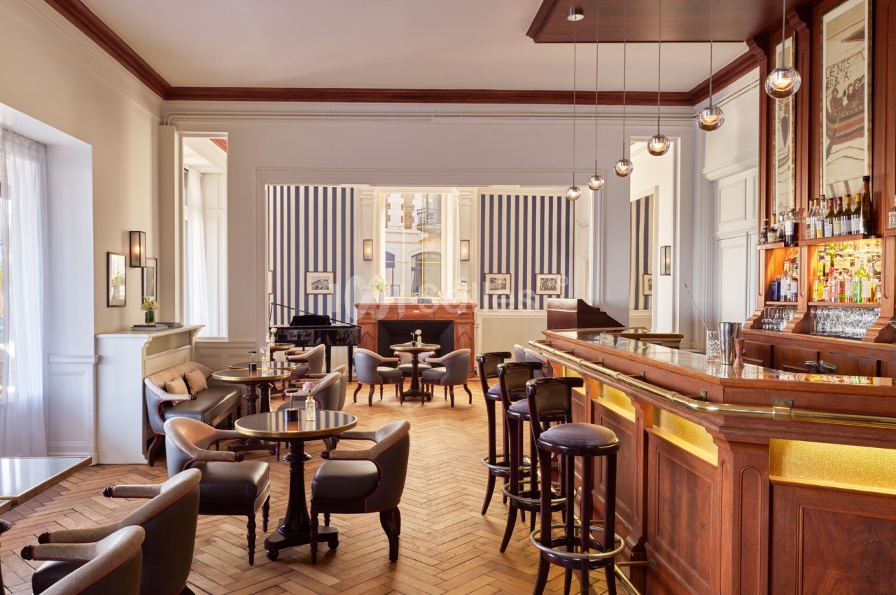 Bar élégant avec comptoir en bois, tables rondes, fauteuils confortables et éclairage suspendu dans un espace lumineux.