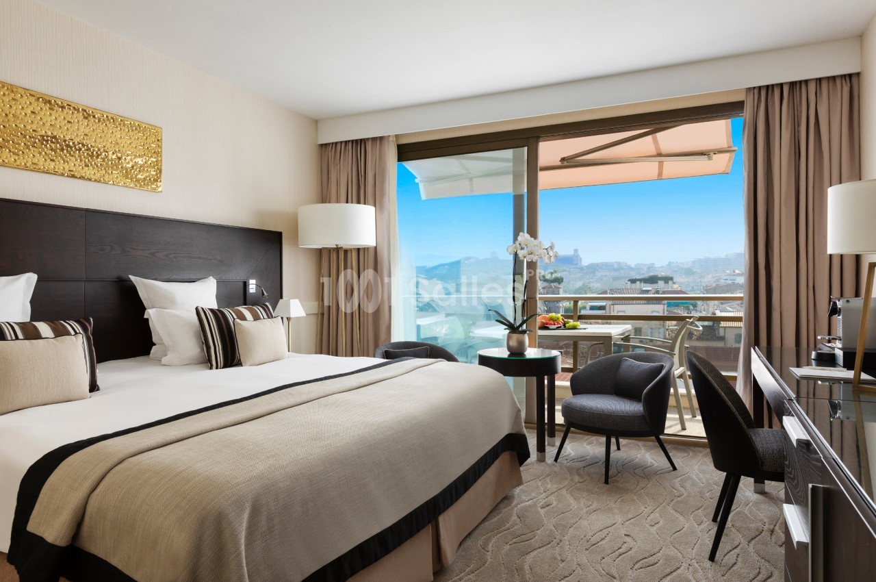 Chambre d'hôtel lumineuse avec lit double, balcon aménagé et vue sur la ville sous un ciel bleu.
