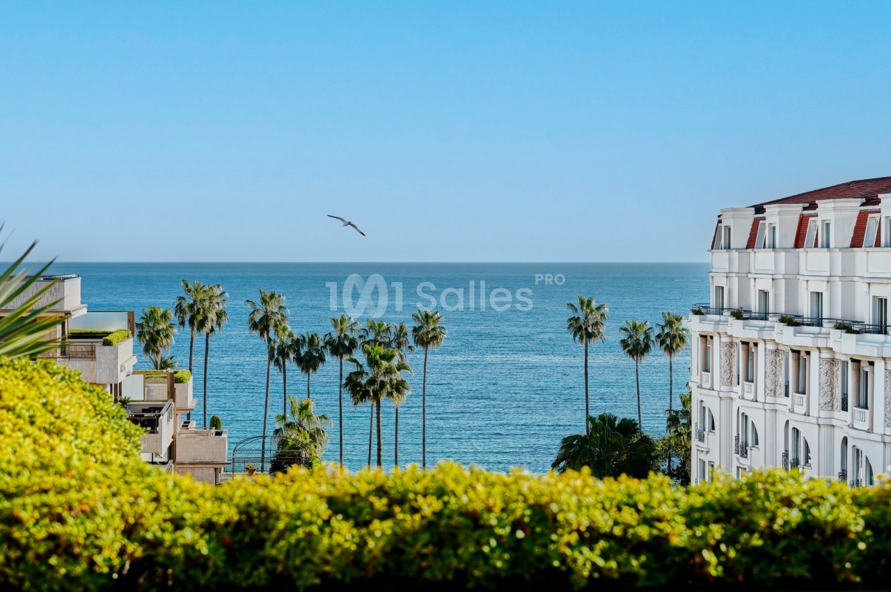 Vue sur la mer avec des palmiers, un bâtiment blanc et un ciel dégagé sous une lumière ensoleillée.