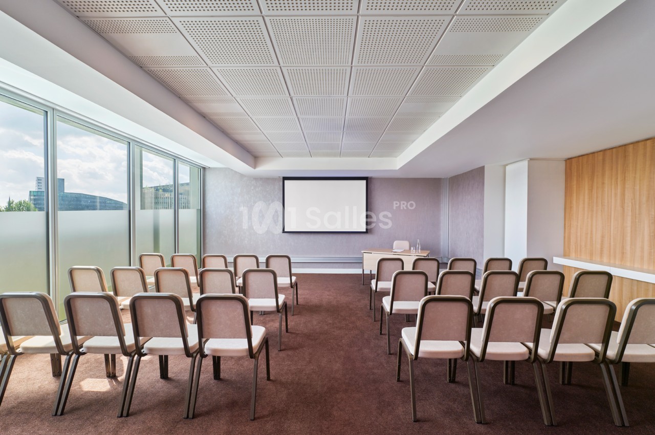 Salle de conférence lumineuse avec rangées de chaises alignées face à un écran de projection.