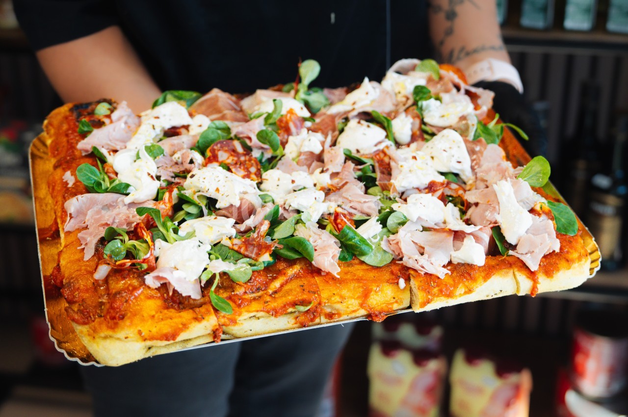 Pizza rectangulaire garnie de jambon, fromage, roquette et tomates séchées, tenue par une personne en arrière-plan flou.