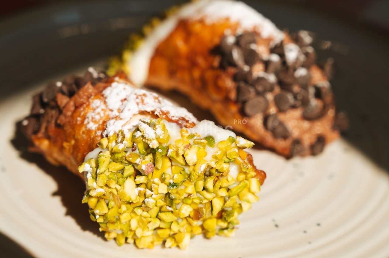 Deux cannoli garnis de crème, l'un décoré de pistaches hachées, l'autre de pépites de chocolat, sur une assiette.