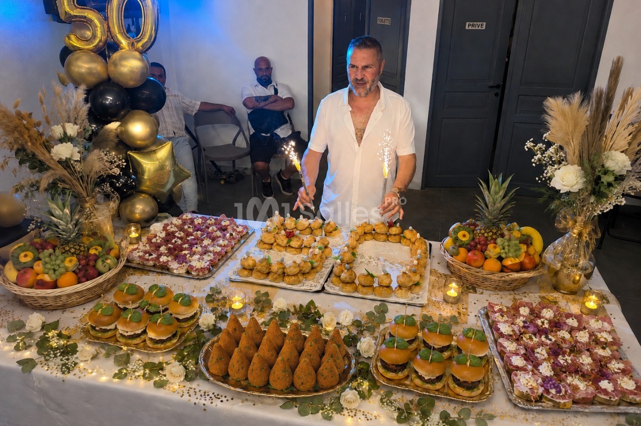 Un homme souriant devant une table garnie de plats variés et décorée pour une fête avec des ballons marquant ’50’.