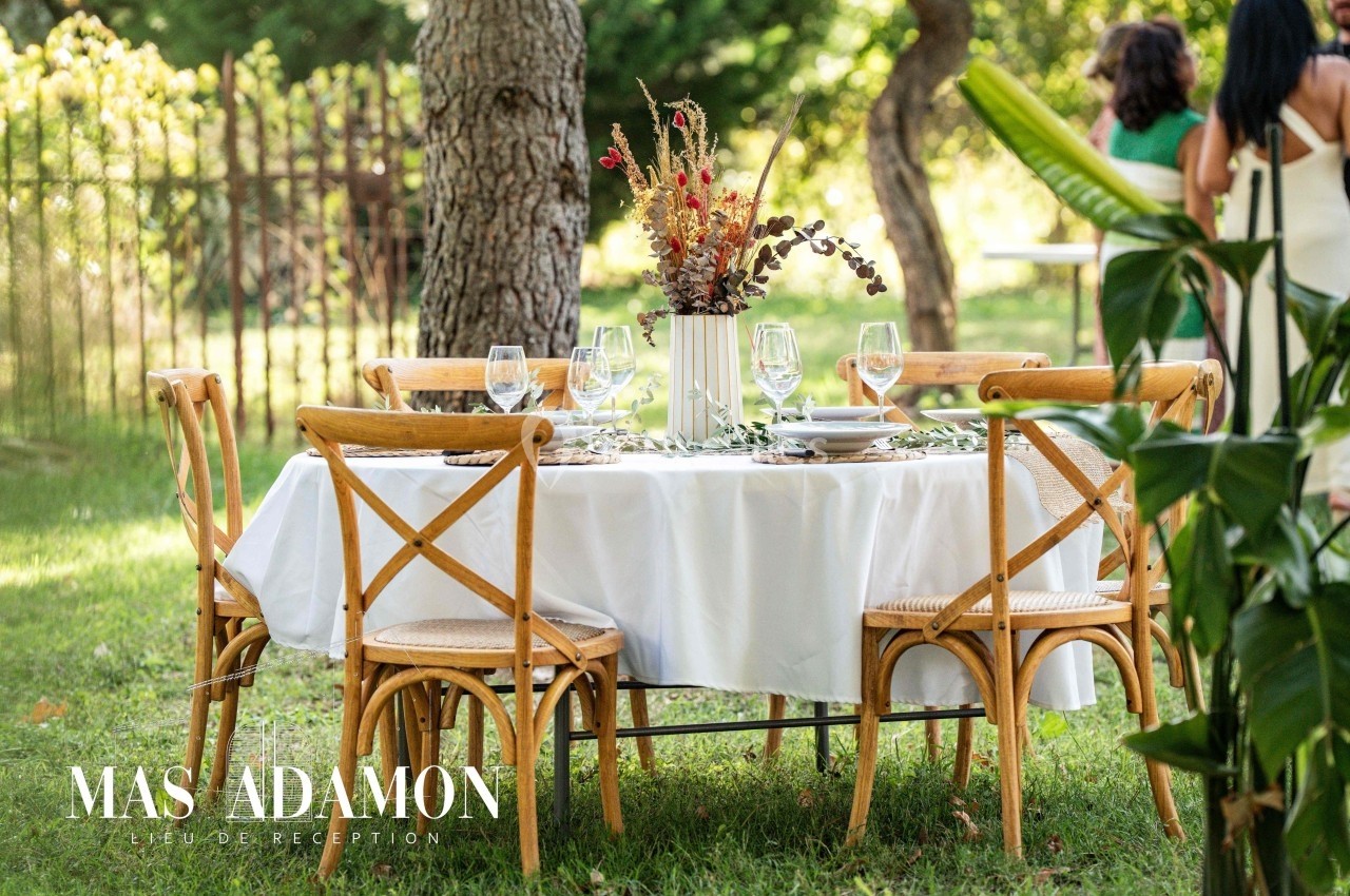Table ronde dressée avec une nappe blanche, chaises en bois et décoration florale, installée dans un jardin verdoyant.