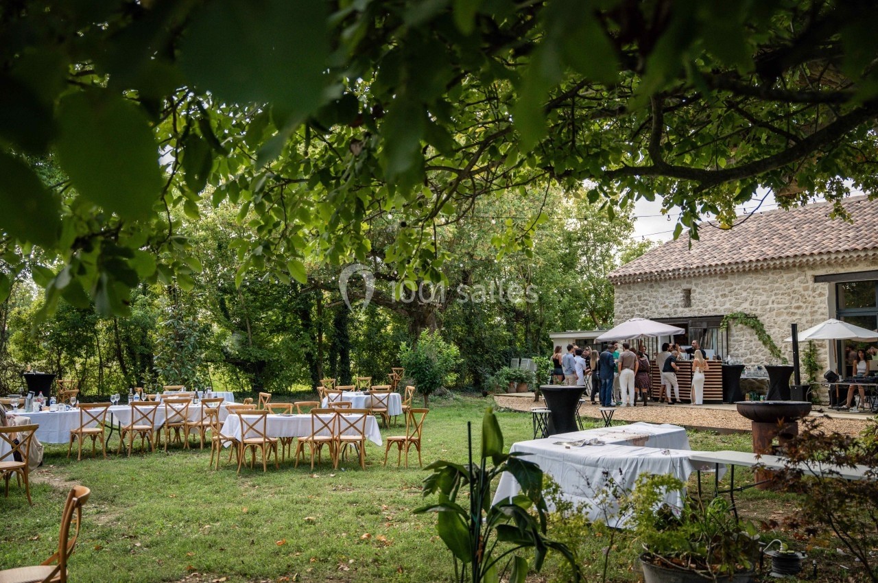 Tables dressées en extérieur sous des arbres, avec des invités rassemblés près d'un bâtiment en pierre.