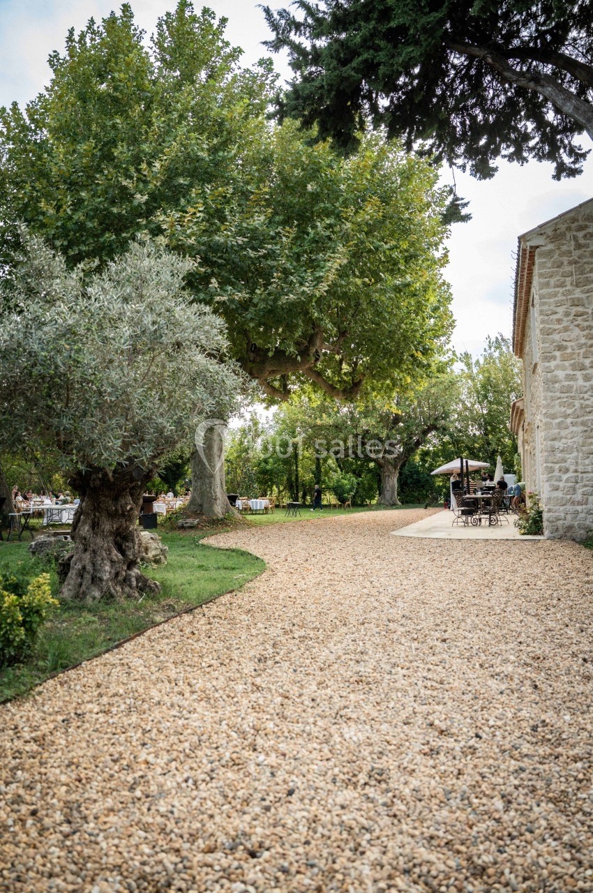 Allée en gravier bordée d'arbres et d'un bâtiment en pierre, menant à une terrasse avec tables et chaises.