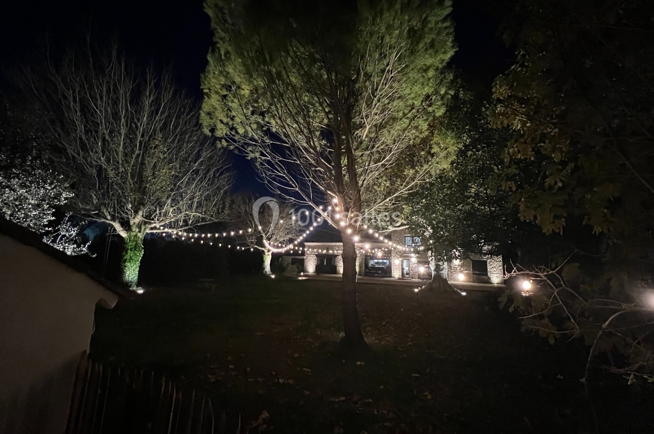 Maison illuminée par des guirlandes lumineuses dans un jardin arboré, vue de nuit.
