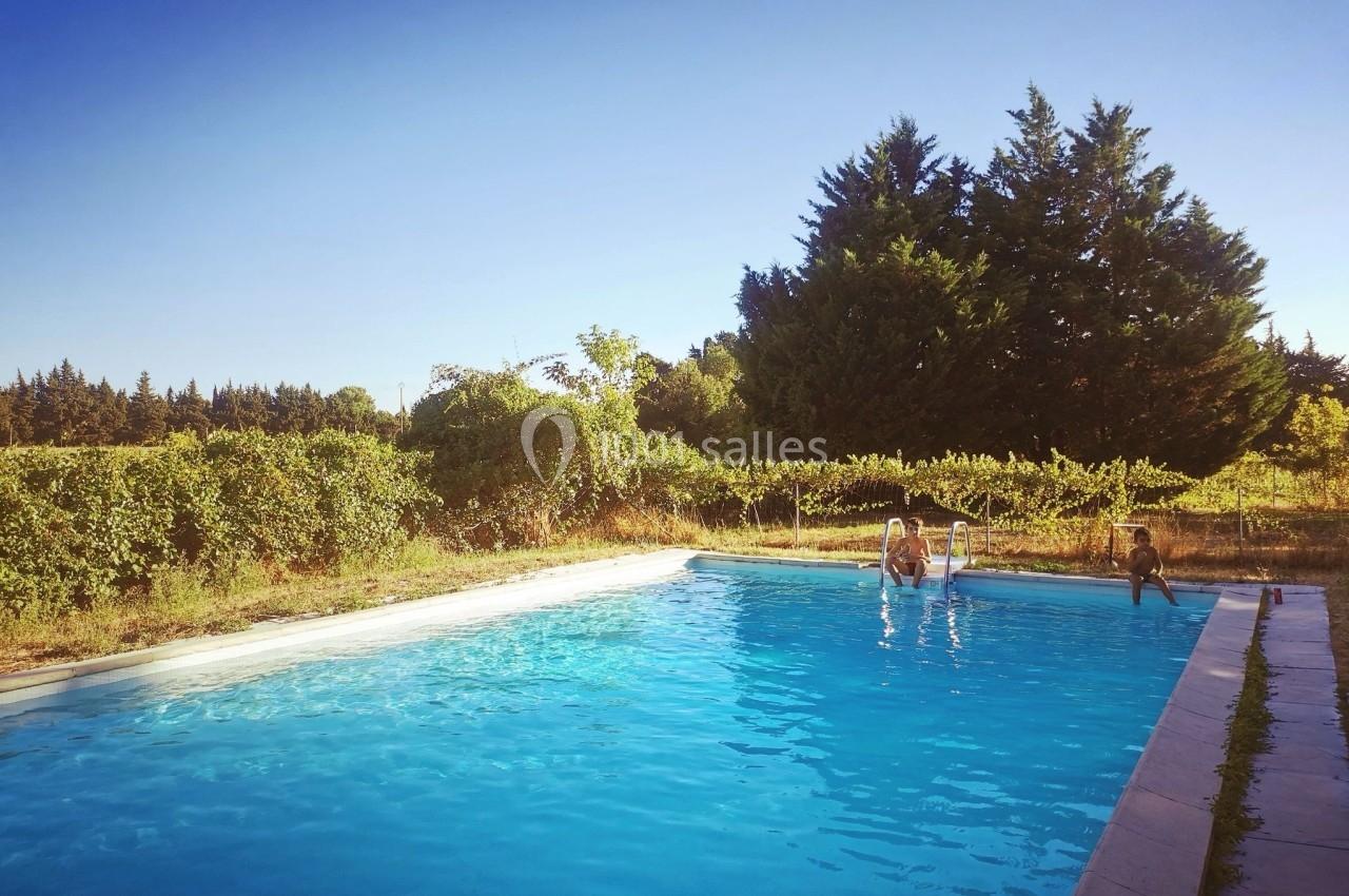 Piscine extérieure entourée de végétation, avec trois personnes assises au bord sous un ciel dégagé.