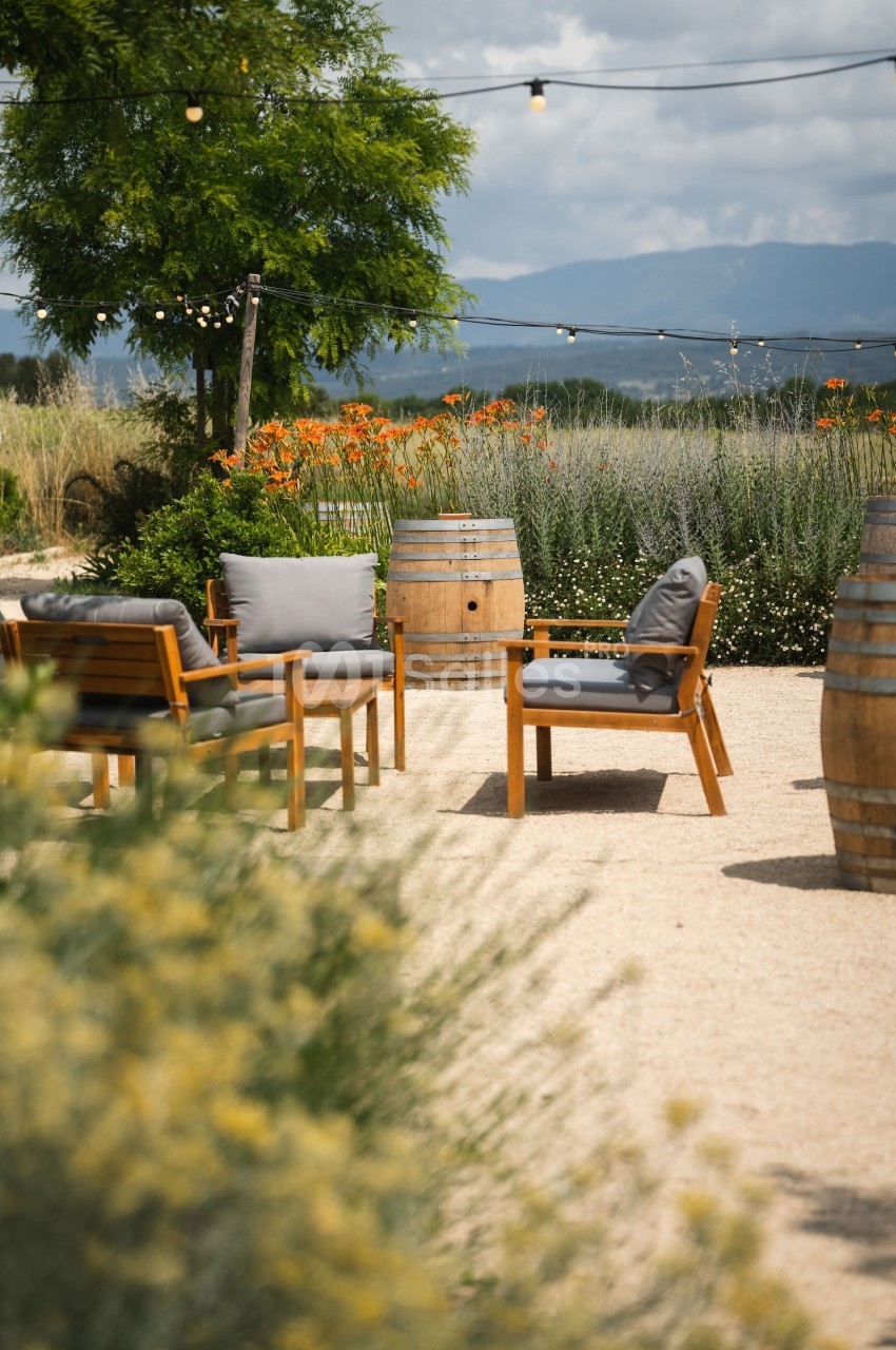 Mobilier de jardin en bois avec coussins gris, entouré de barriques, fleurs et végétation sous un ciel nuageux.