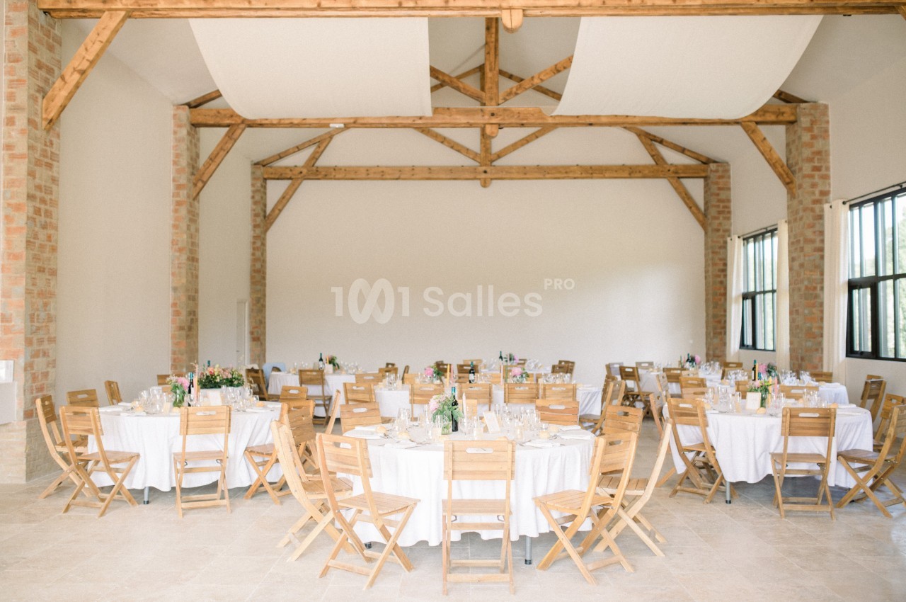 Salle lumineuse avec des tables rondes dressées, entourées de chaises en bois, sous une charpente apparente.