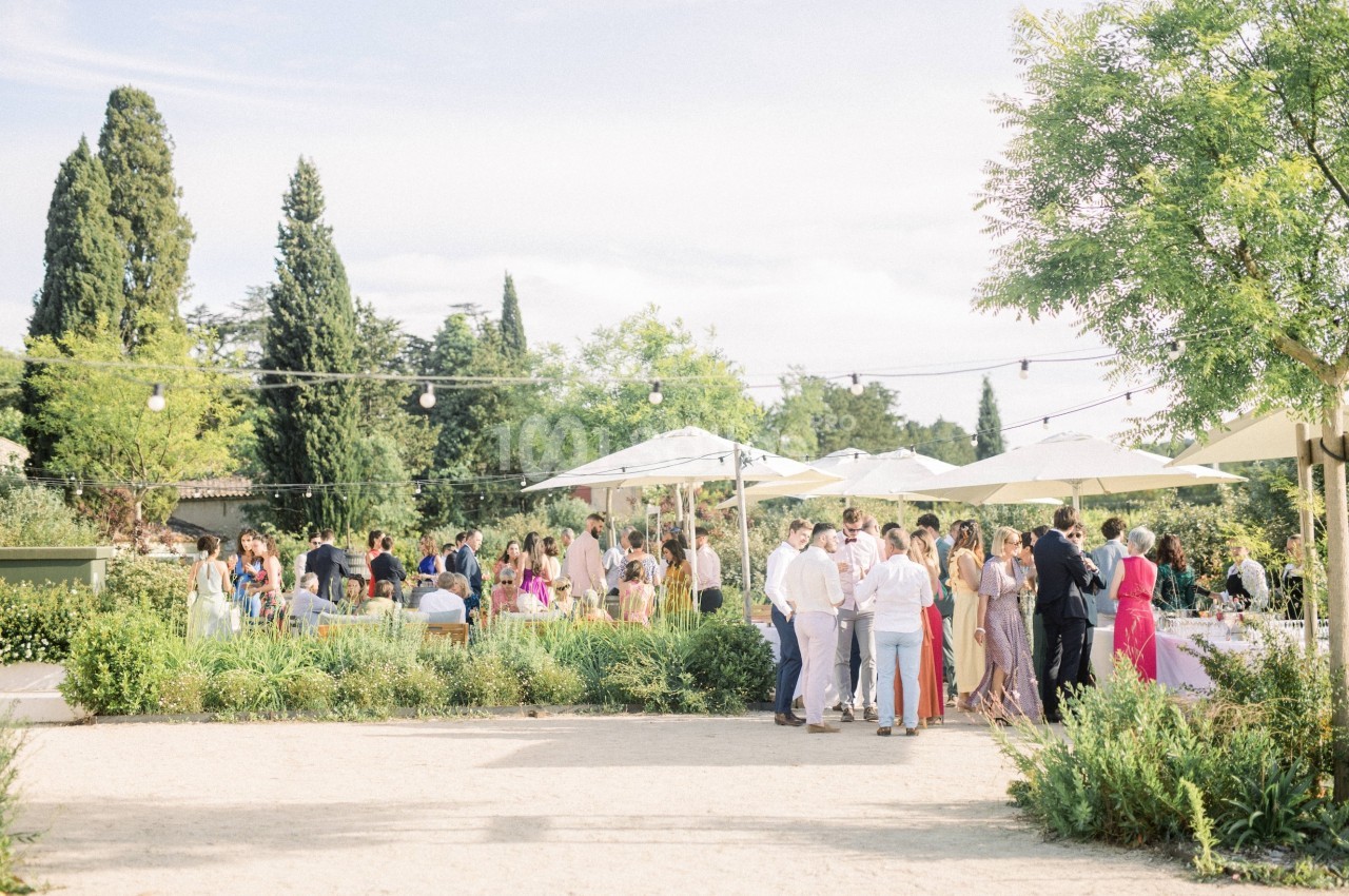 Groupe de personnes rassemblées dans un jardin ensoleillé avec parasols, arbres et guirlandes lumineuses en arrière-plan.