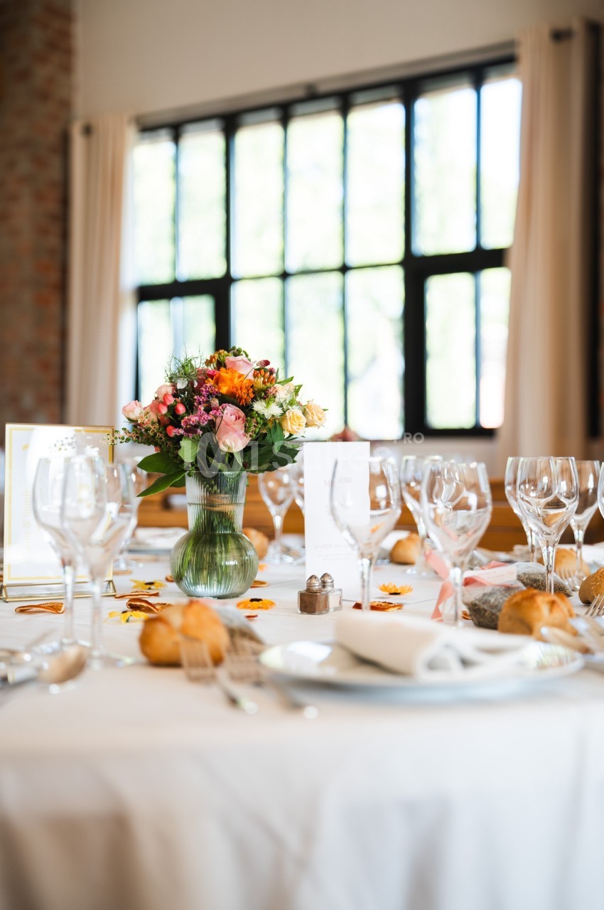 Table dressée avec nappes blanches, verres, couverts, bouquet de fleurs colorées et lumière naturelle en arrière-plan.