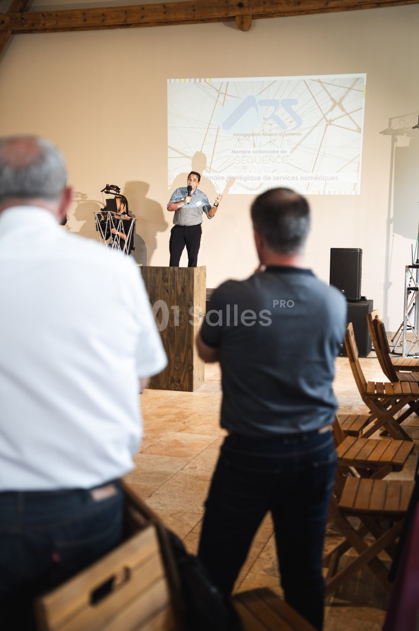 Une personne présente sur scène devant un public dans une salle avec projection d'une diapositive en arrière-plan.