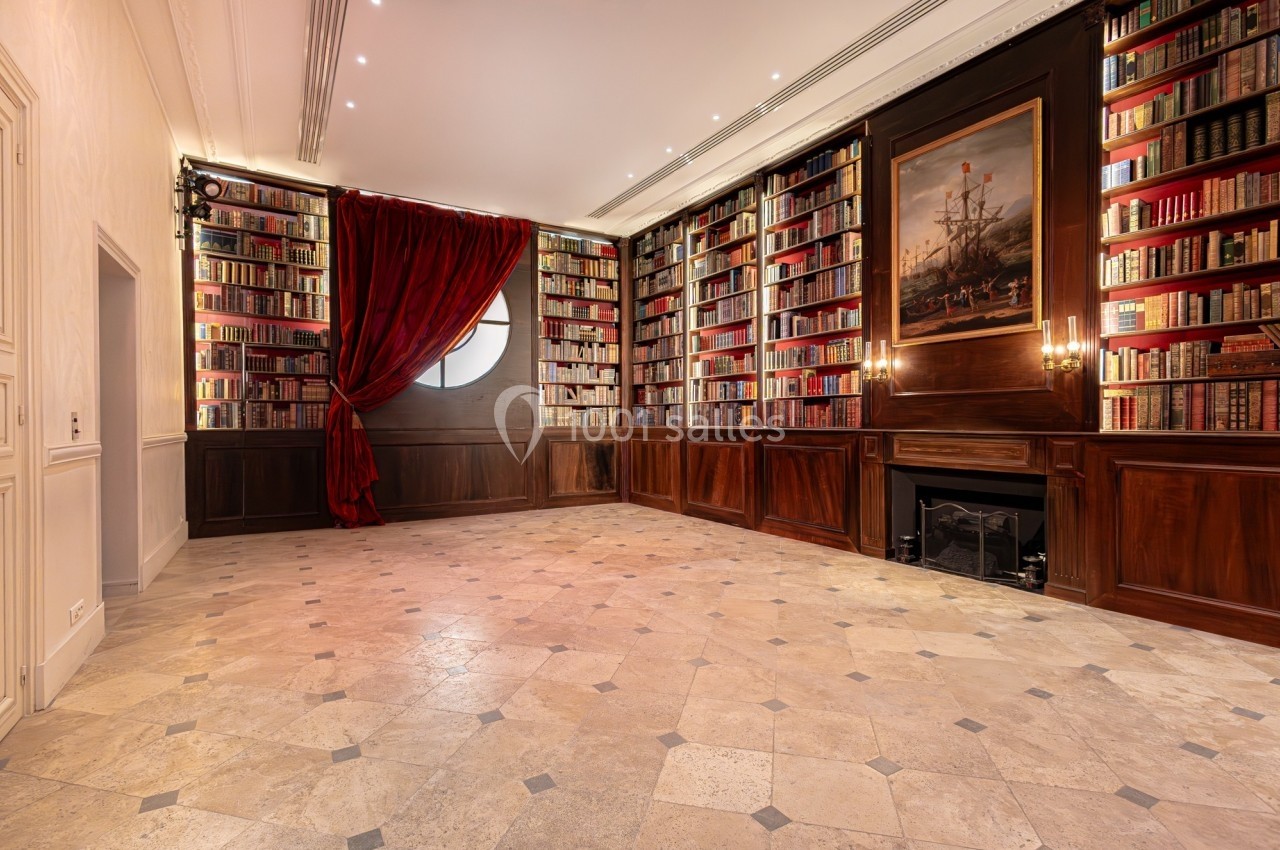 Bibliothèque élégante avec murs couverts de livres, cheminée, tableau, rideaux rouges et sol en pierre claire.