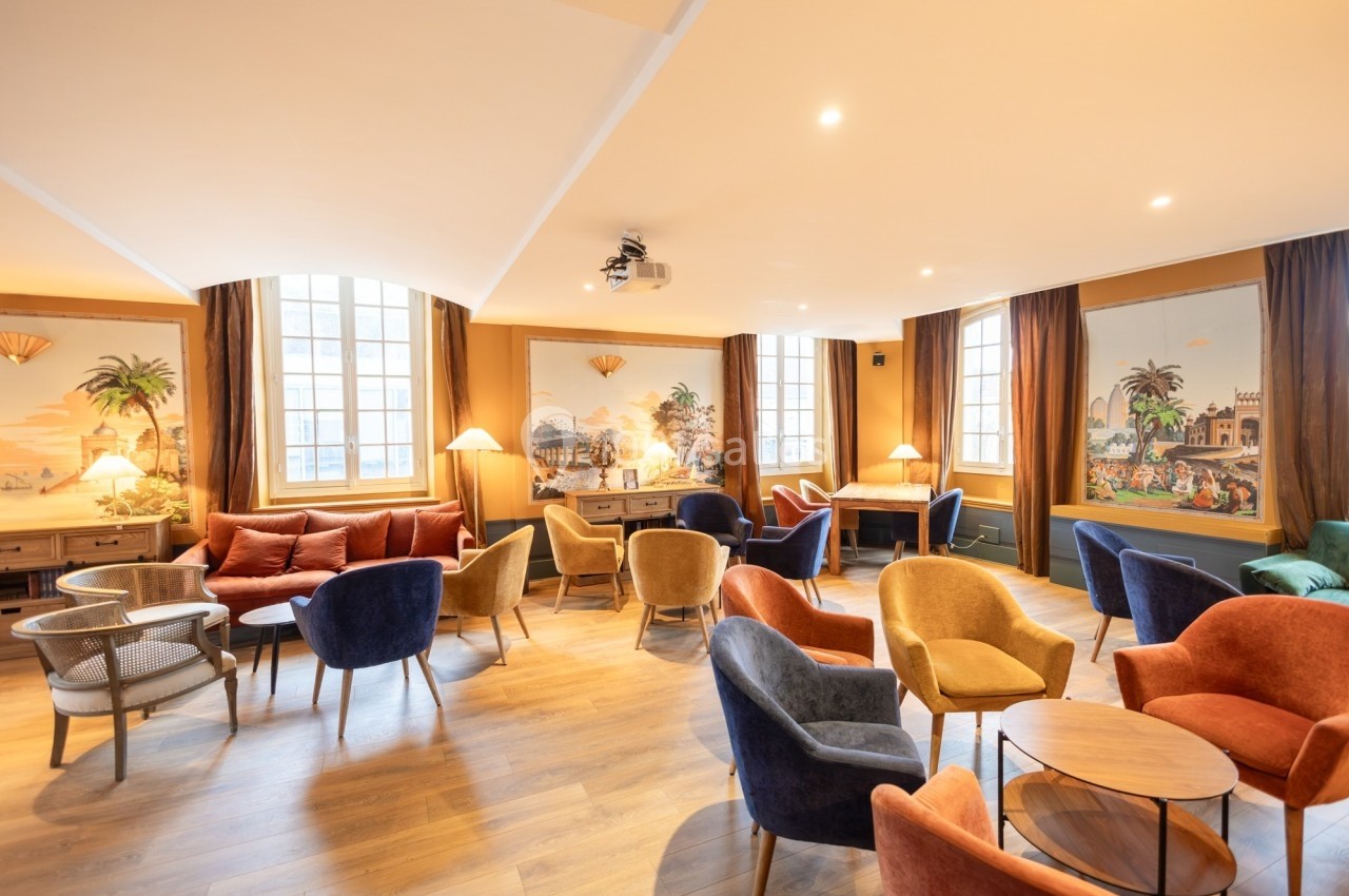 Salle lumineuse avec fauteuils colorés, tables basses, parquet en bois et fresques murales décoratives.