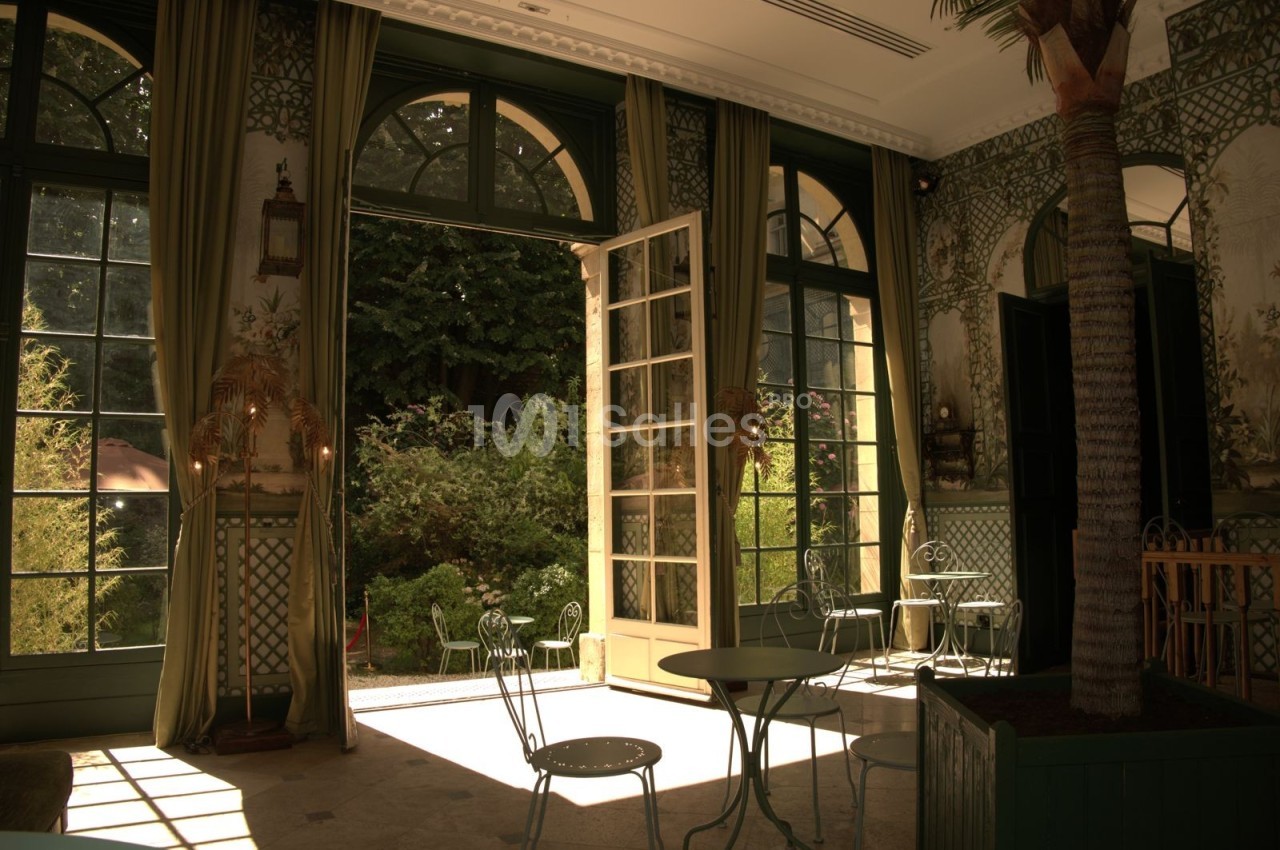 Salle lumineuse avec grandes baies vitrées ouvertes sur un jardin verdoyant, mobilier en métal et décorations végétales.