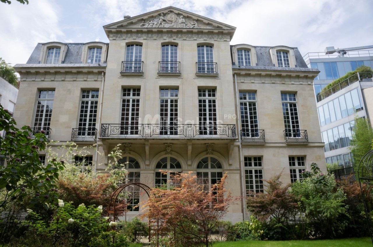 Façade d'un bâtiment ancien en pierre avec grandes fenêtres, entouré de végétation et d'arbustes colorés.