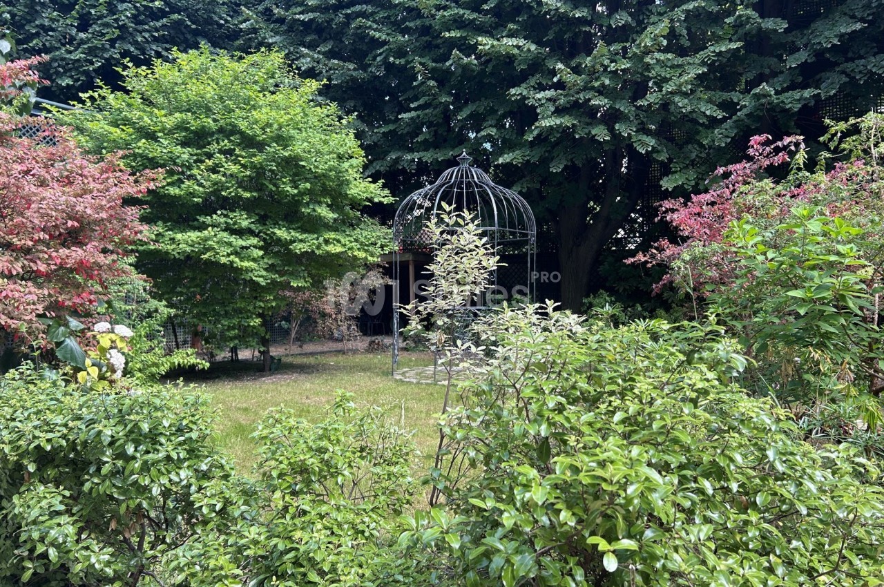 Vue d'un jardin verdoyant avec des arbres, des buissons et une grande cage métallique décorative au centre.