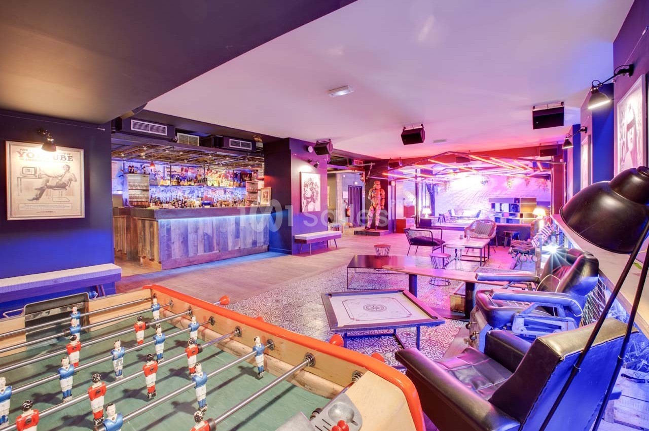 Salle de loisirs avec baby-foot, bar éclairé, fauteuils et ambiance lumineuse colorée.