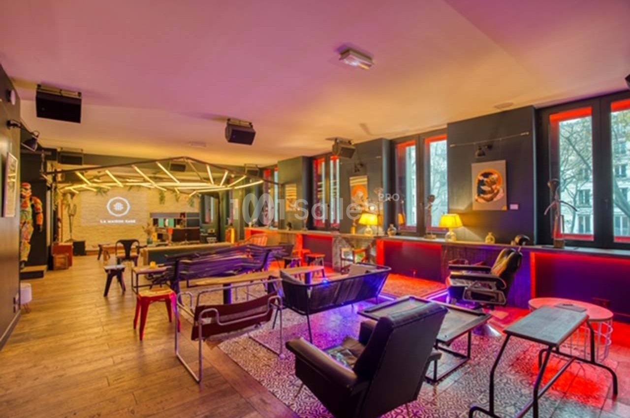 Salle moderne avec mobilier éclectique, éclairage coloré et grandes fenêtres donnant sur l'extérieur.