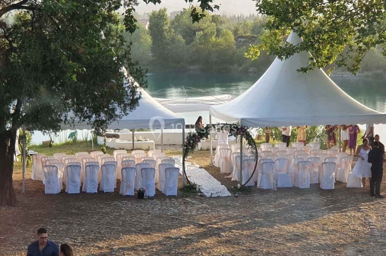 Chaises blanches alignées sous des tentes blanches près d'un lac, entourées d'arbres, préparées pour une cérémonie en plein…