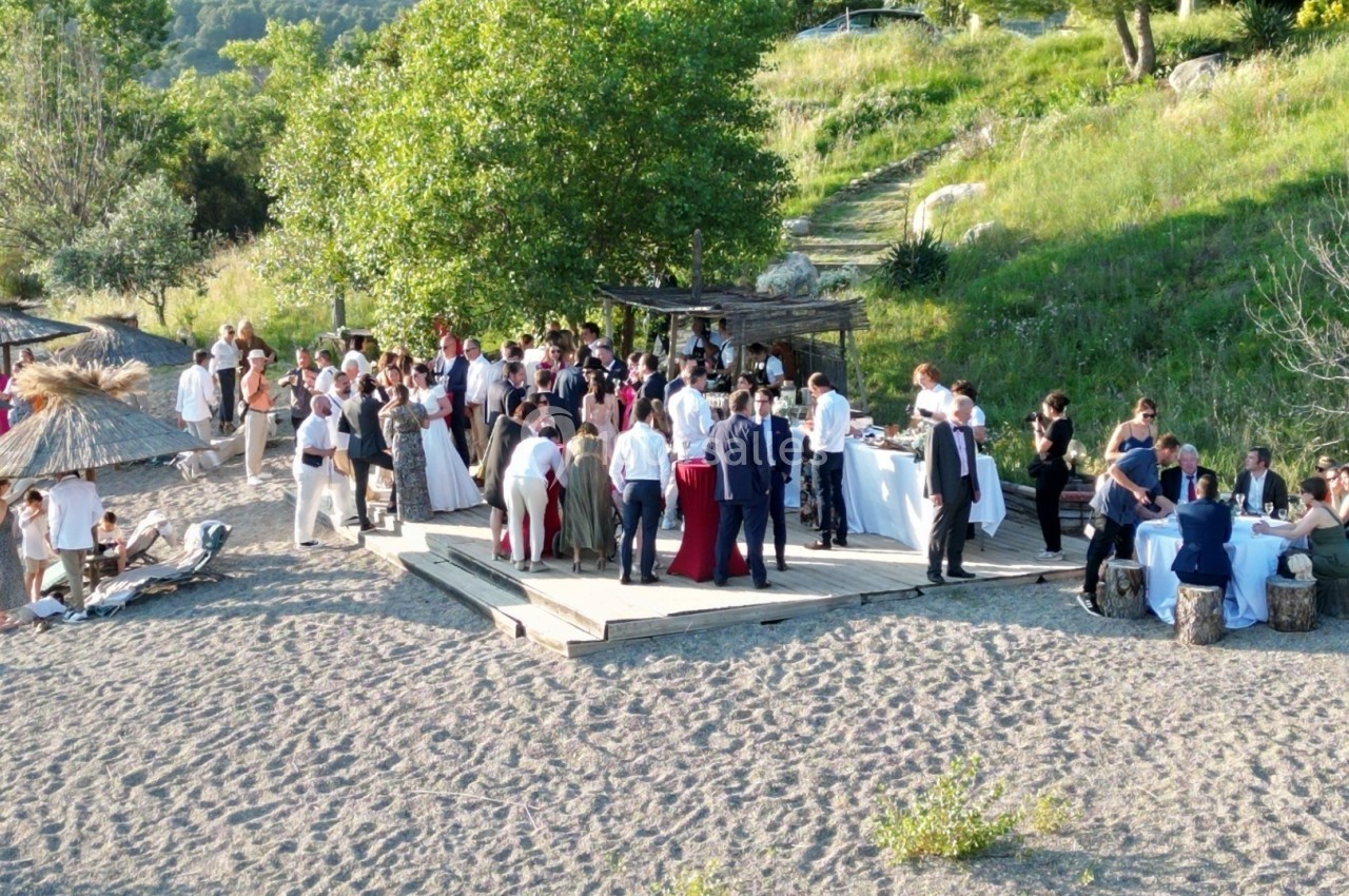 Groupe de personnes rassemblées sur une terrasse en bois au bord d'une plage, entourée de verdure.