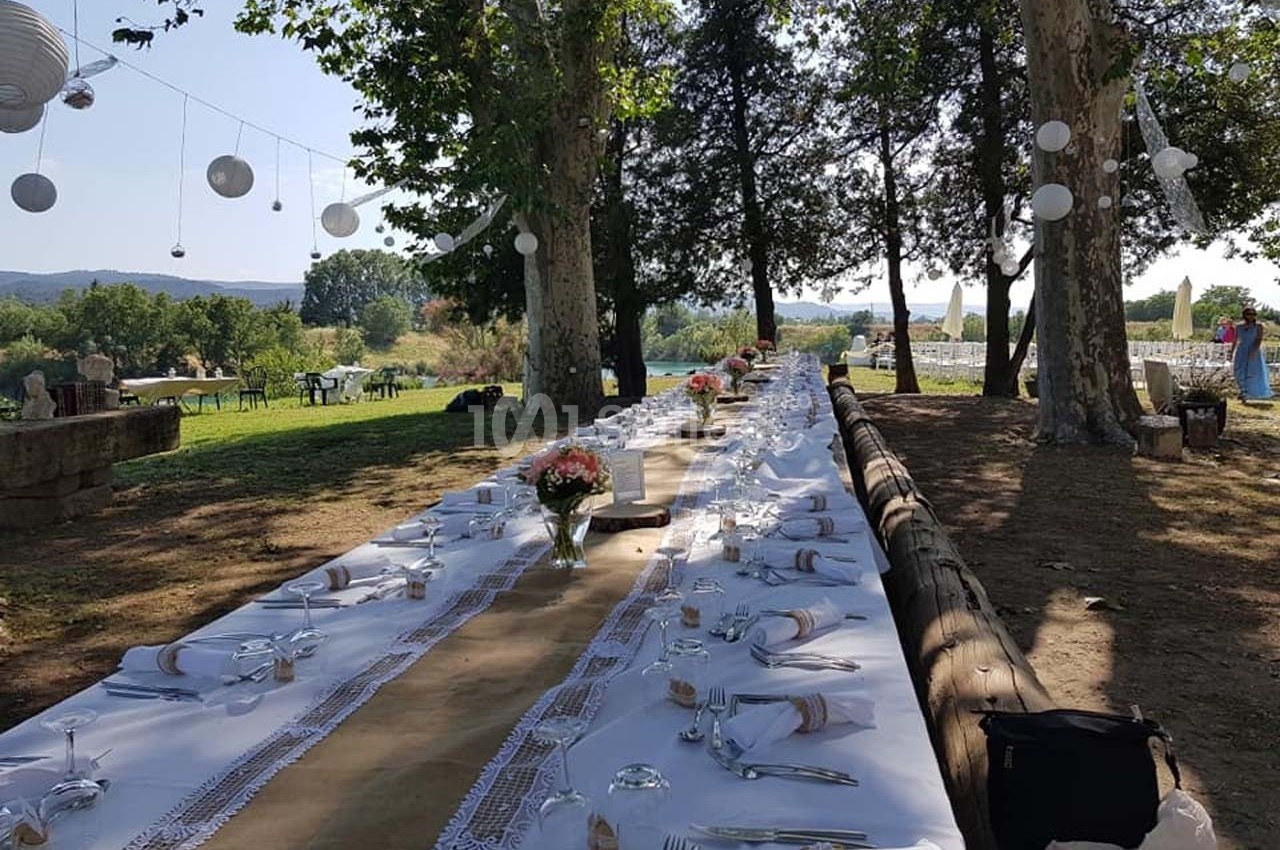 Grande table extérieure décorée pour un repas, sous des arbres avec des lanternes suspendues, dans un cadre champêtre.
