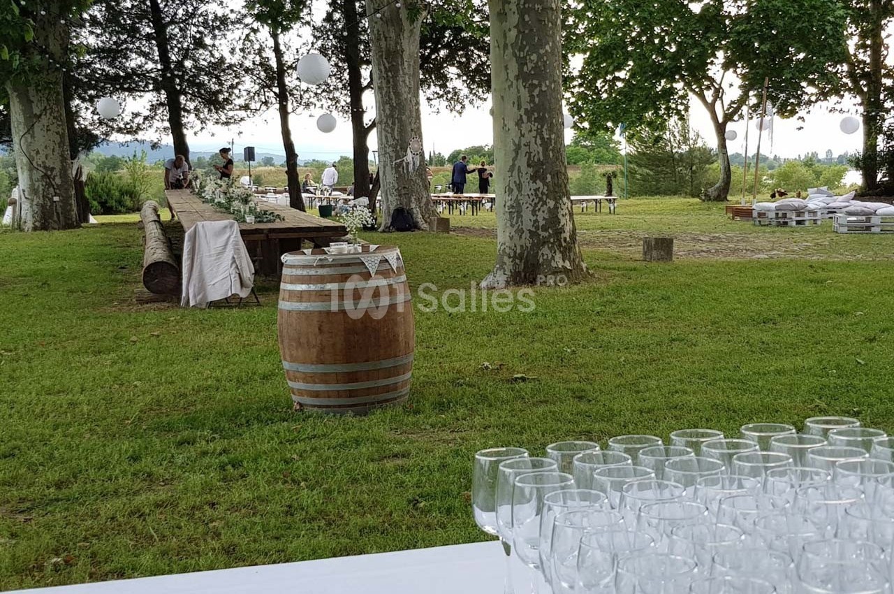 Table dressée avec des verres à pied en premier plan, espace extérieur verdoyant avec tables et décorations en arrière-plan.