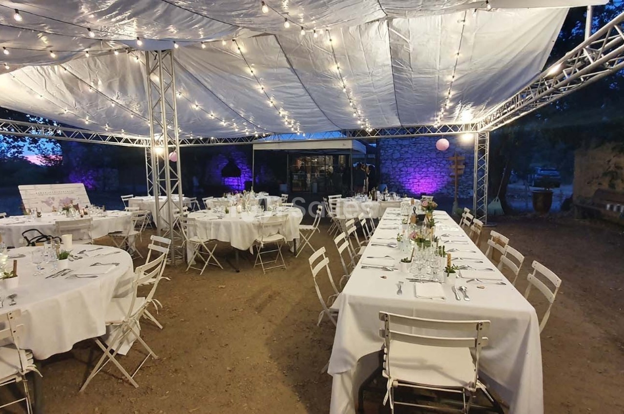 Tables dressées avec nappes blanches sous une tente éclairée de guirlandes lumineuses, dans un cadre extérieur en soirée.