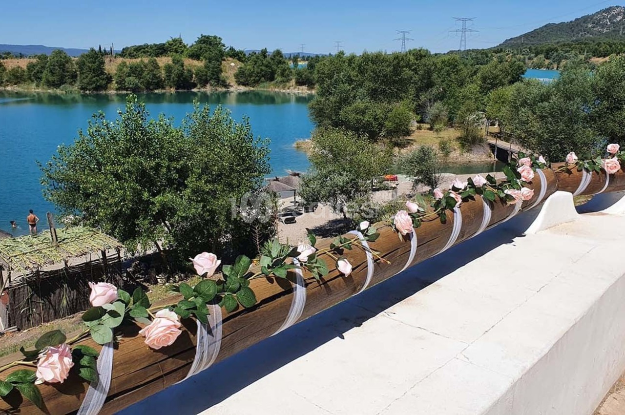Barrière en bois décorée de roses artificielles roses, surplombant un lac entouré de végétation et de collines.