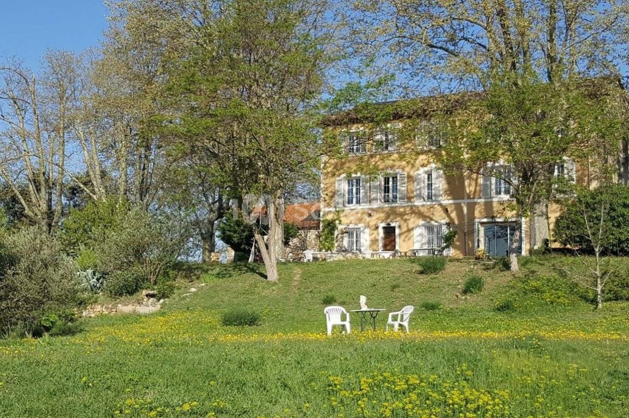 Maison en pierre entourée d'arbres et d'une pelouse fleurie, avec deux chaises blanches et une table au premier plan.