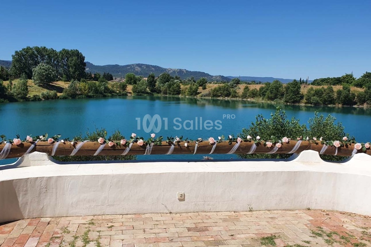 Vue sur un lac entouré de verdure, avec une balustrade décorée de fleurs et rubans sous un ciel dégagé.