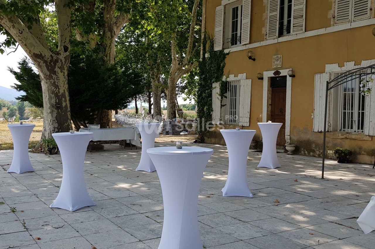 Terrasse pavée avec des tables hautes recouvertes de housses blanches, devant une bâtisse ancienne entourée d'arbres.