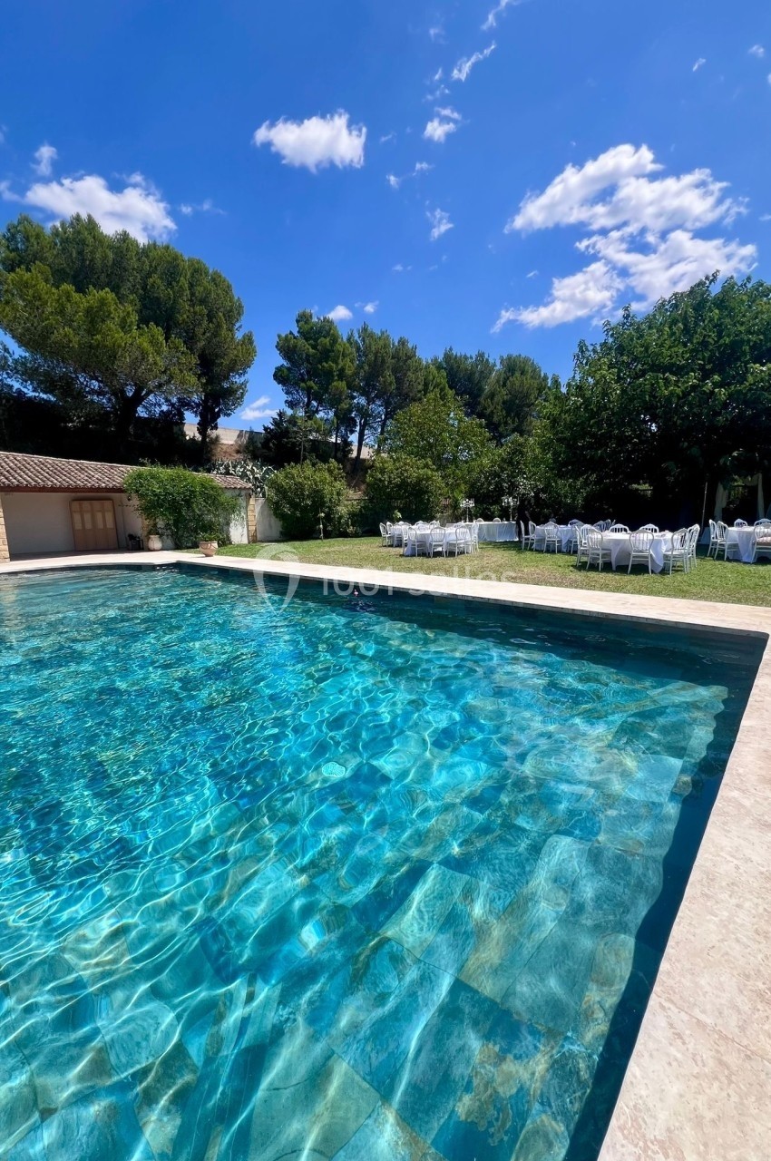 Piscine extérieure entourée de verdure, avec des tables blanches dressées sous des arbres par une journée ensoleillée.