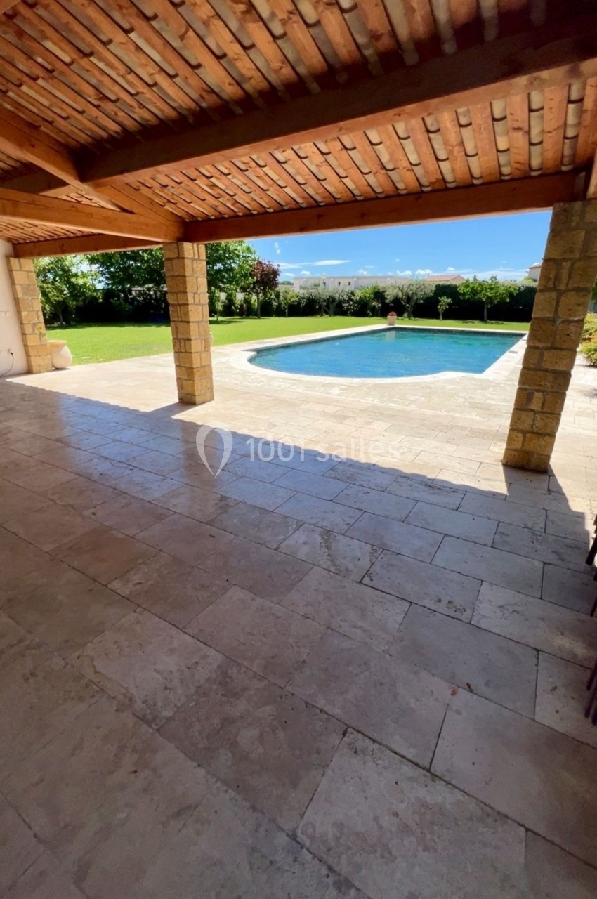 Vue depuis une terrasse couverte en pierre donnant sur une piscine entourée de verdure et d'un ciel dégagé.