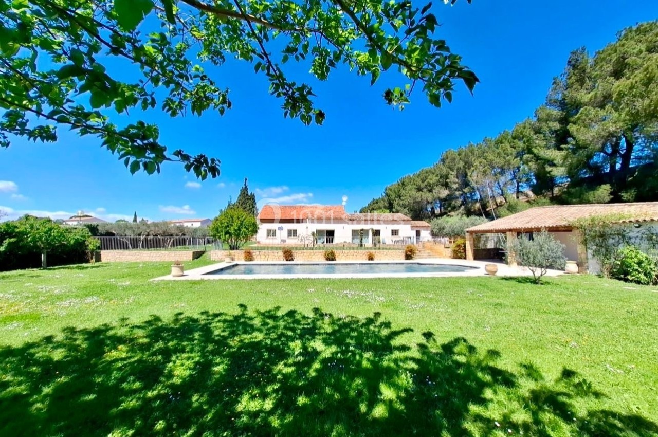 Maison blanche avec toit en tuiles, piscine et grand jardin verdoyant, entourée d'arbres sous un ciel bleu.