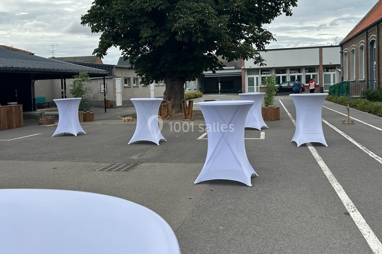 Tables hautes blanches disposées en extérieur sur un parking, avec un arbre et des bâtiments en arrière-plan.