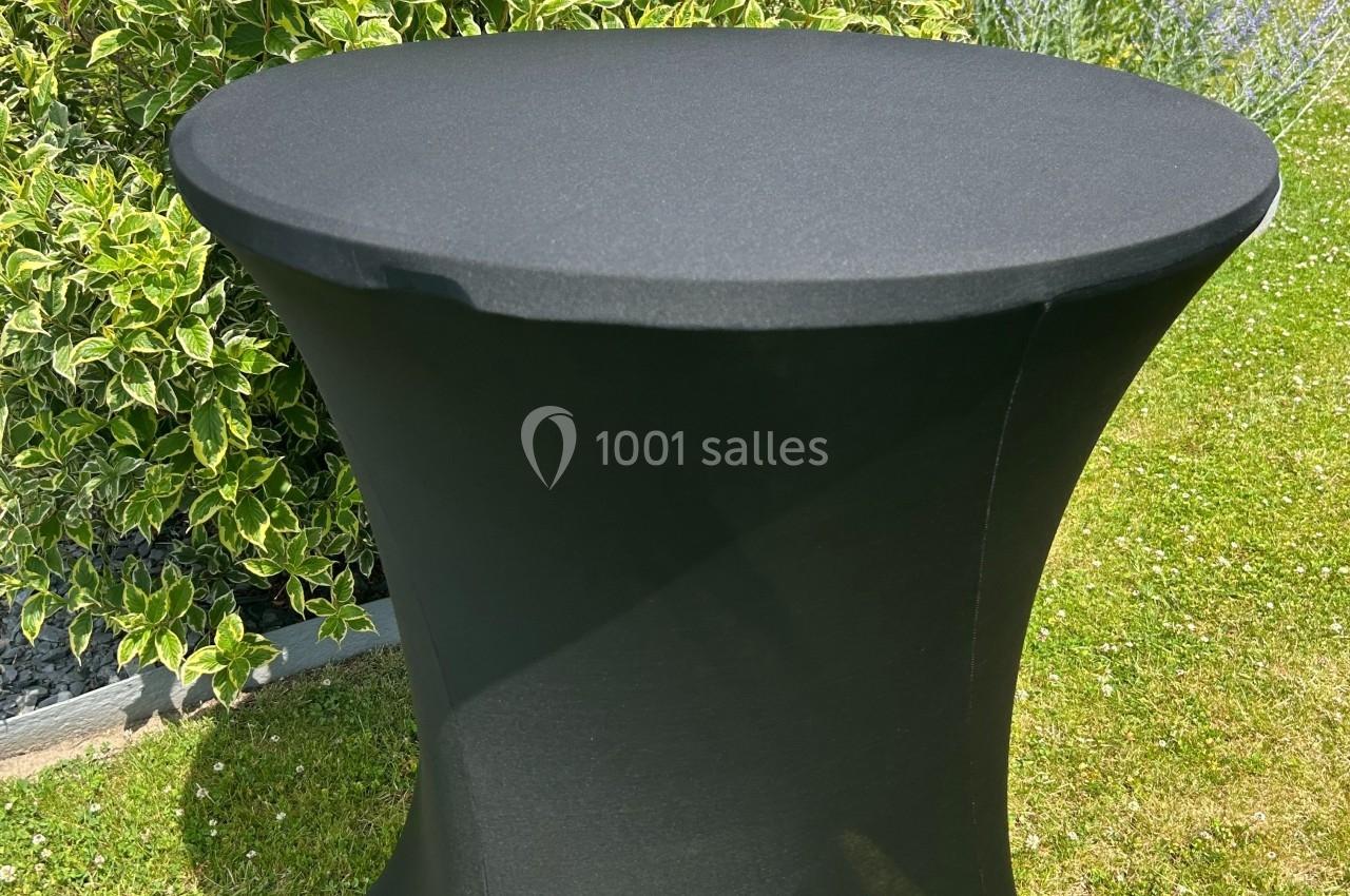 Table haute recouverte d'une housse noire, placée sur une pelouse avec des buissons en arrière-plan.