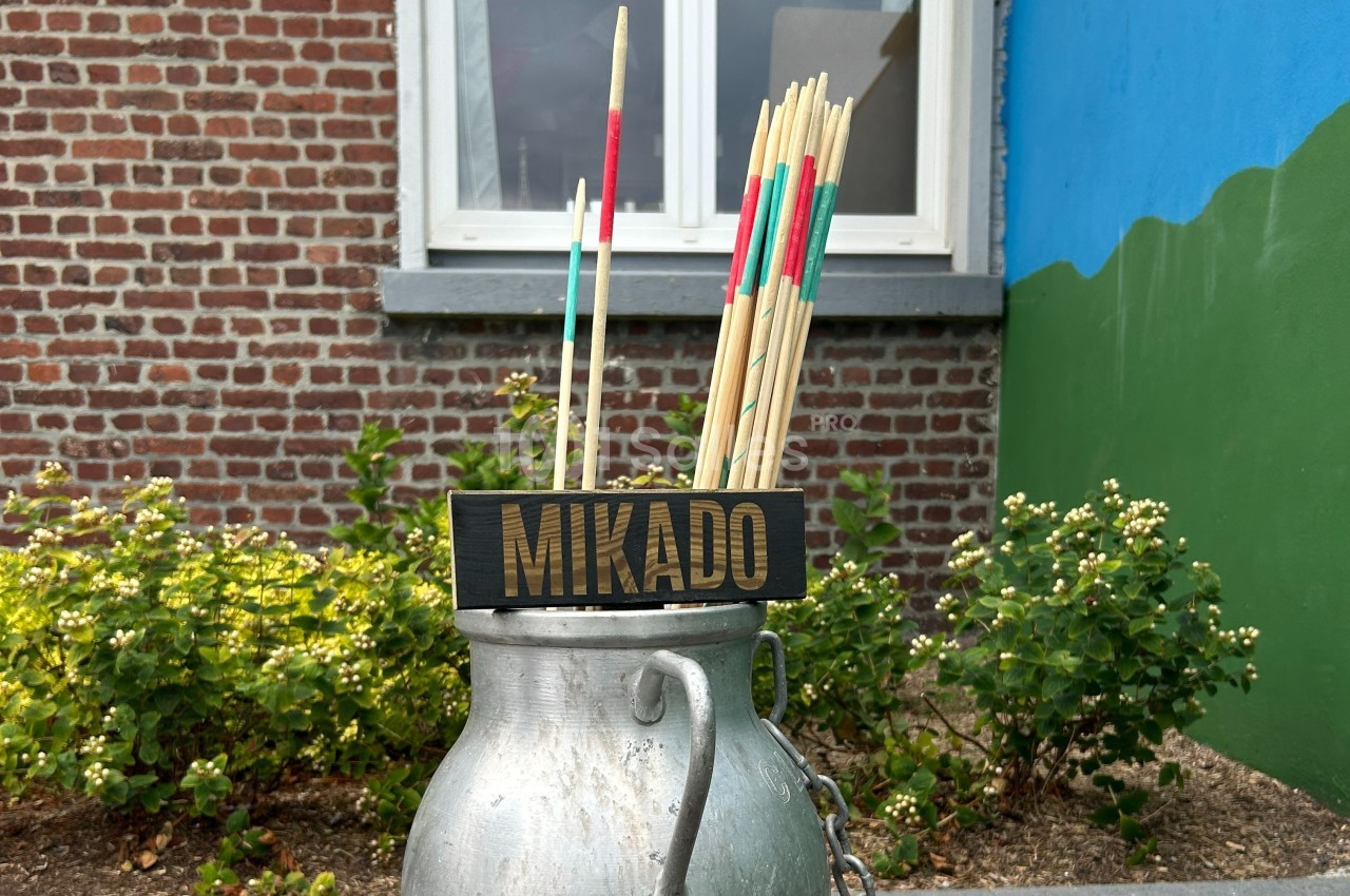 Des bâtons de mikado géants dans un pot en métal devant un mur de briques et un jardin fleuri.