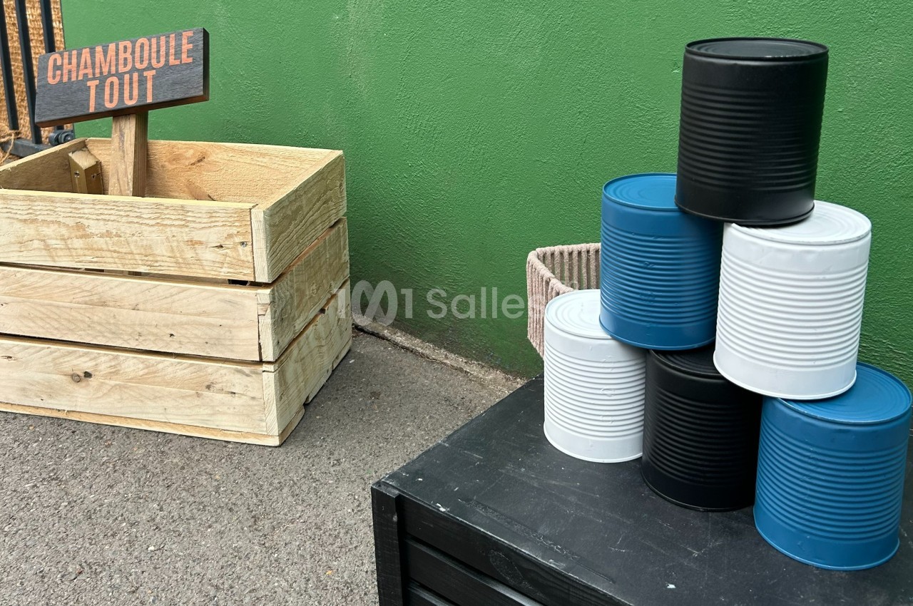 Pyramide de boîtes de conserve colorées posée sur une caisse noire, avec un panneau ’Chamboule Tout’ en arrière-plan.