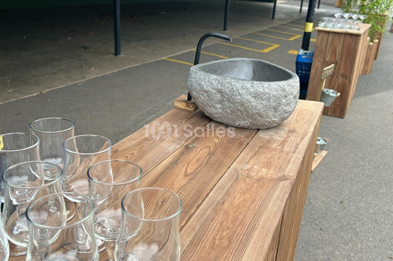 Bol en pierre avec un robinet sur une table en bois, entouré de verres transparents, en extérieur.
