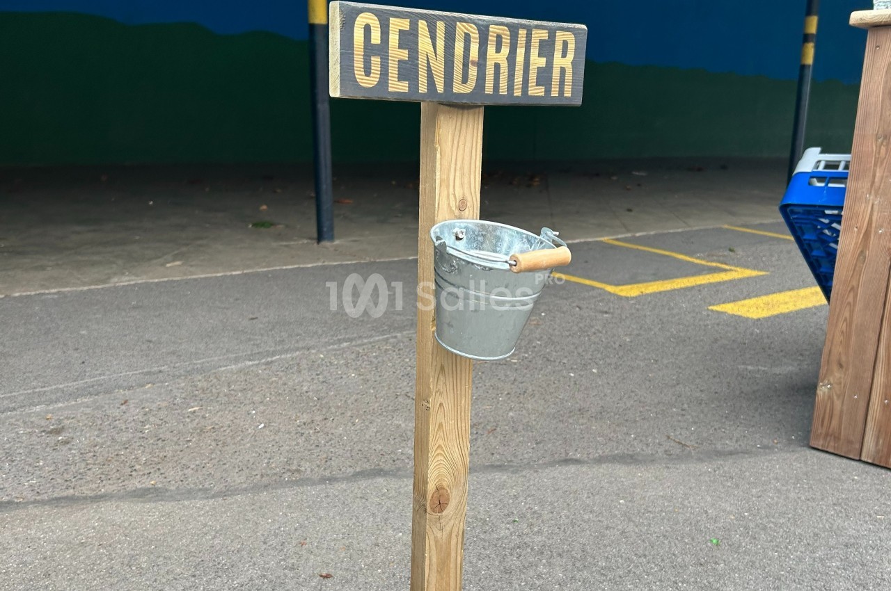 Poteau en bois avec un seau métallique suspendu servant de cendrier, placé sur un sol asphalté.