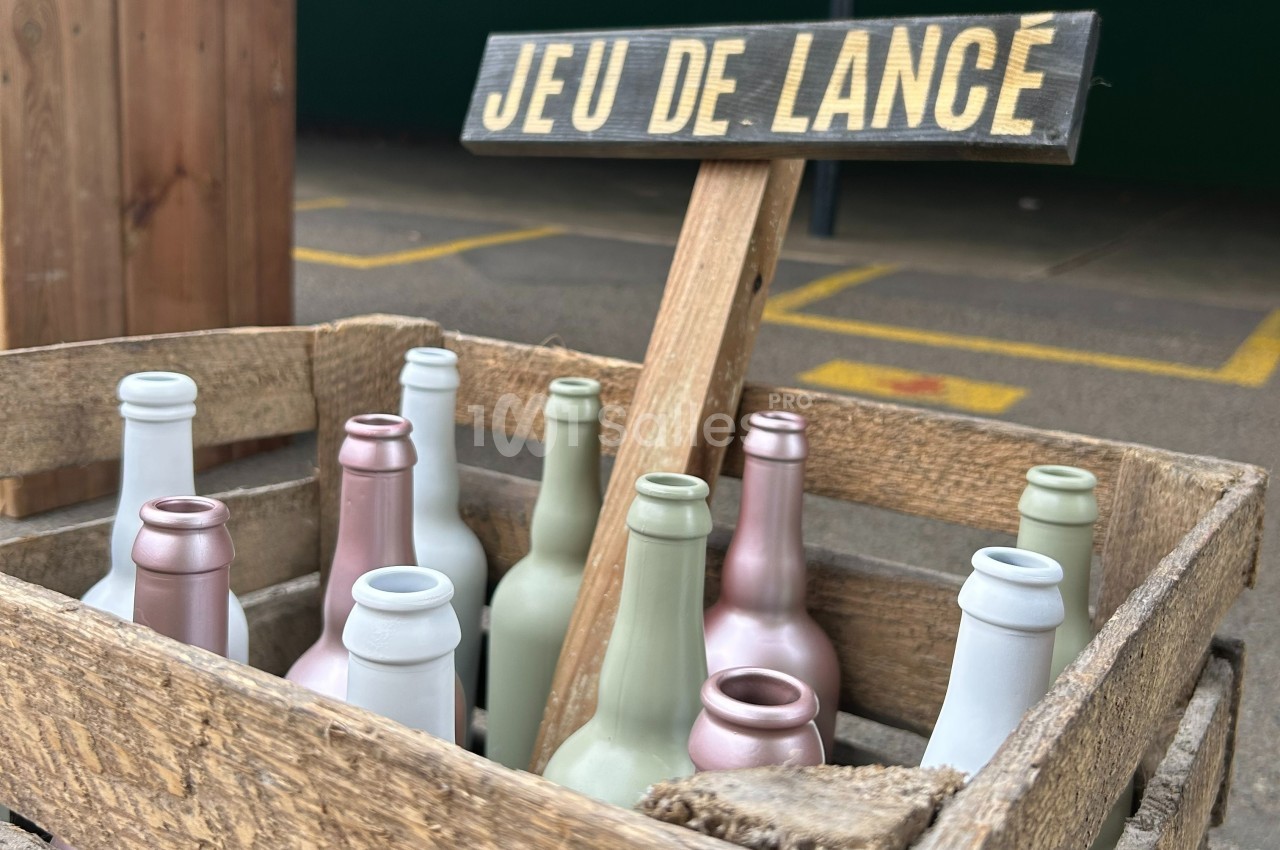 Caisse en bois contenant des bouteilles colorées, avec un panneau indiquant ’Jeu de lance’ en arrière-plan.