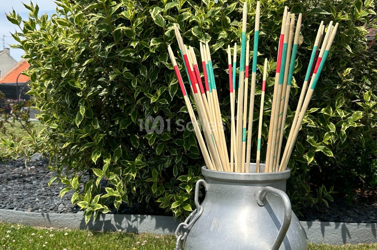 Bâtonnets de mikado géants colorés dans un pot en métal, posés devant un buisson dans un jardin.