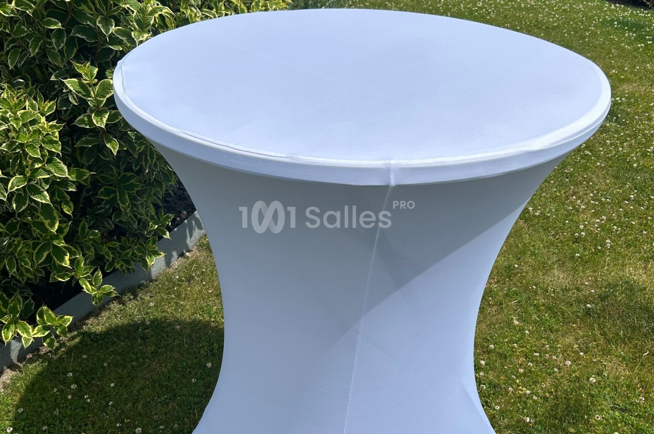 Table ronde recouverte d'une housse blanche, placée sur une pelouse avec des arbustes en arrière-plan.