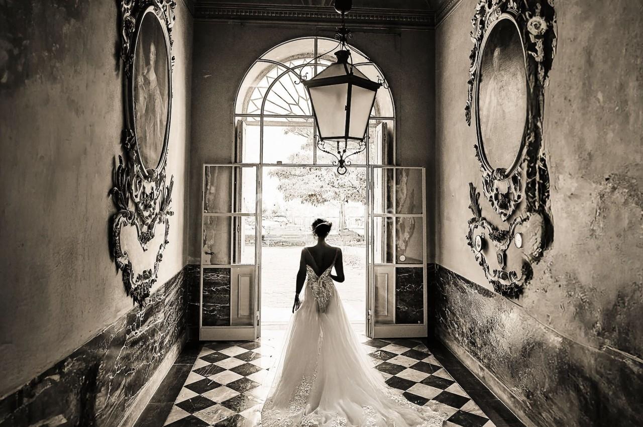 Une mariée en robe blanche marche dans un couloir ancien avec un sol en damier et des fresques murales.