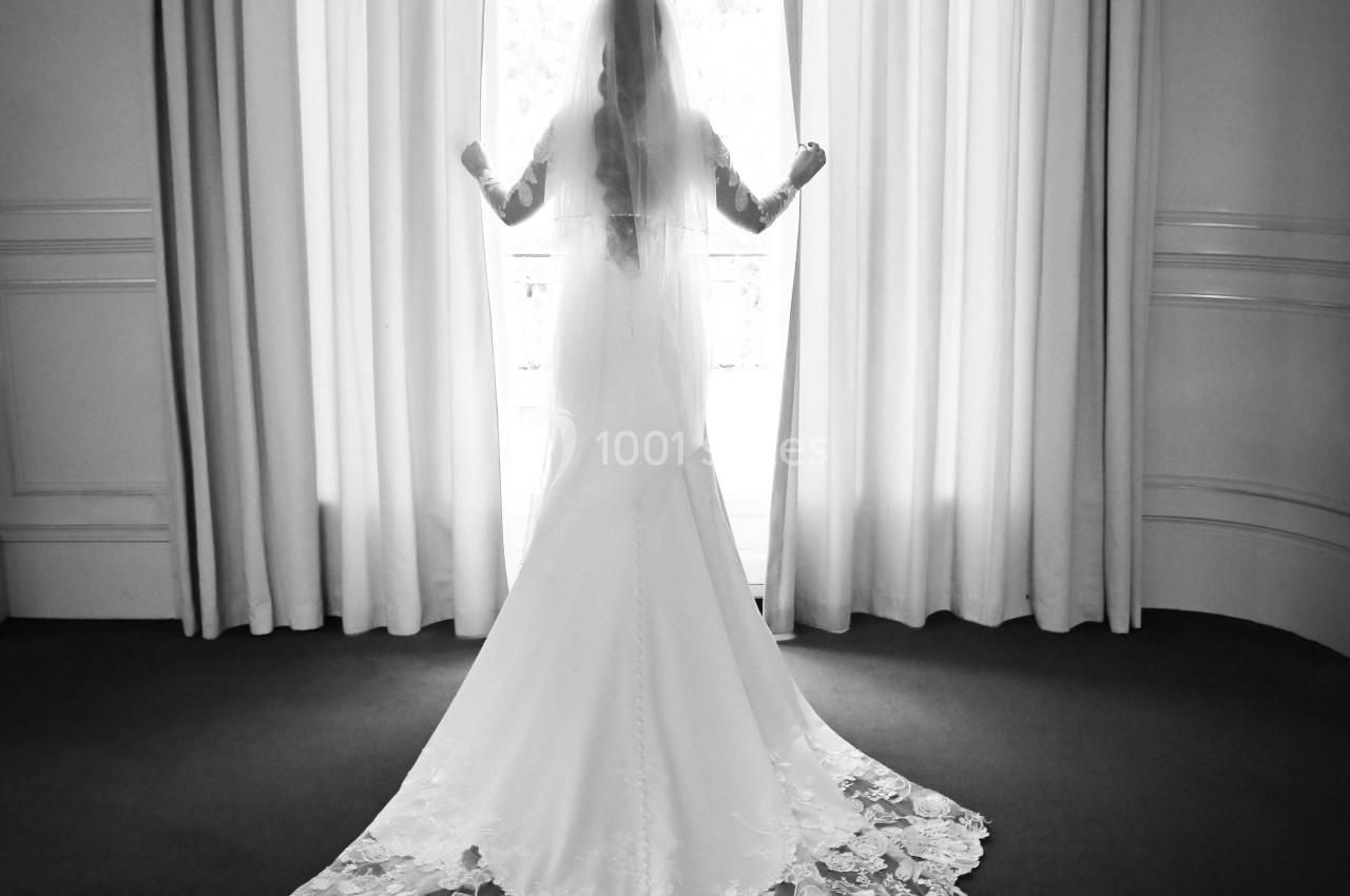 Une mariée en robe blanche avec une longue traîne en dentelle se tient devant une fenêtre lumineuse.