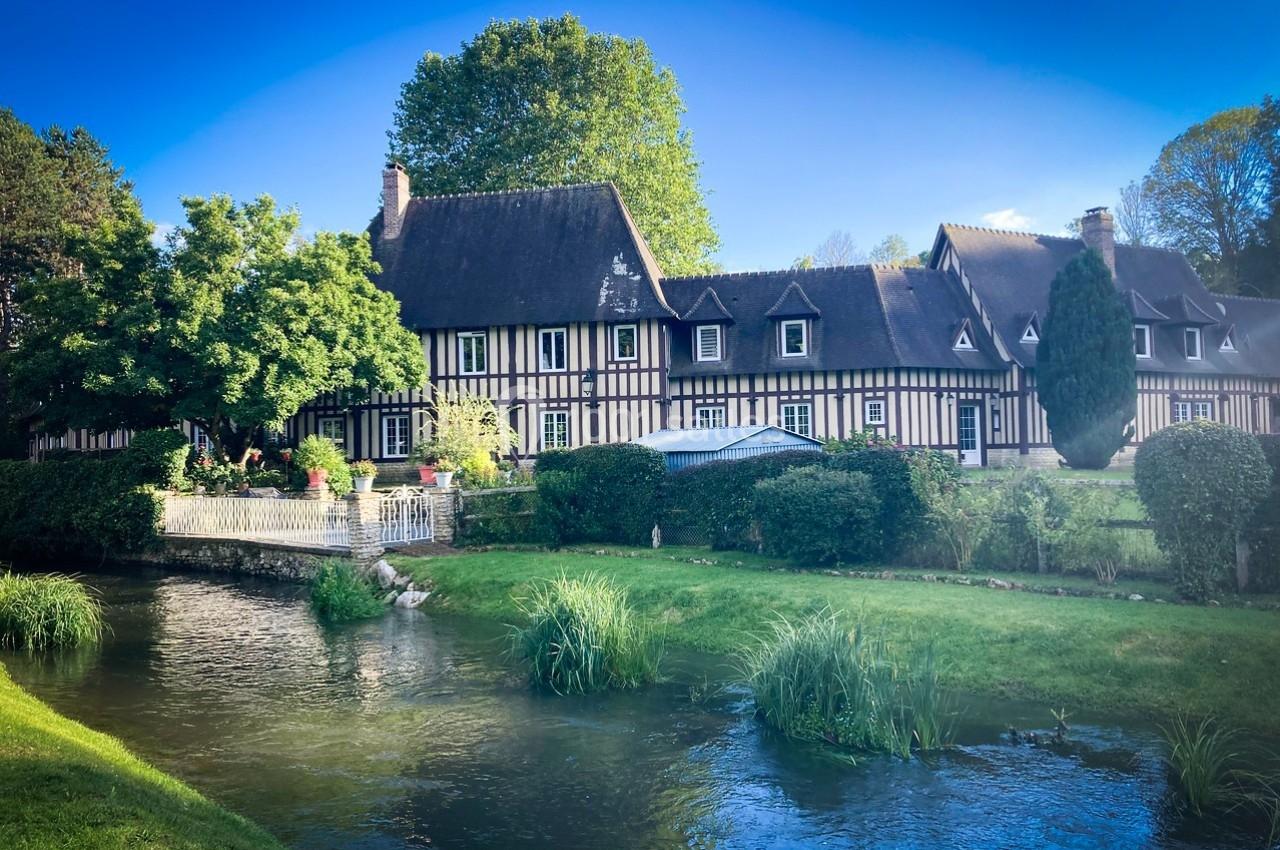 Maison à colombages entourée de verdure, avec un ruisseau au premier plan sous un ciel bleu clair.