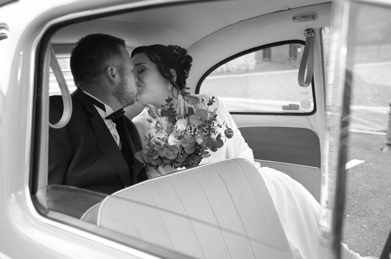Un couple en tenue de mariage s'embrasse à l'intérieur d'une voiture ancienne, la mariée tenant un bouquet.